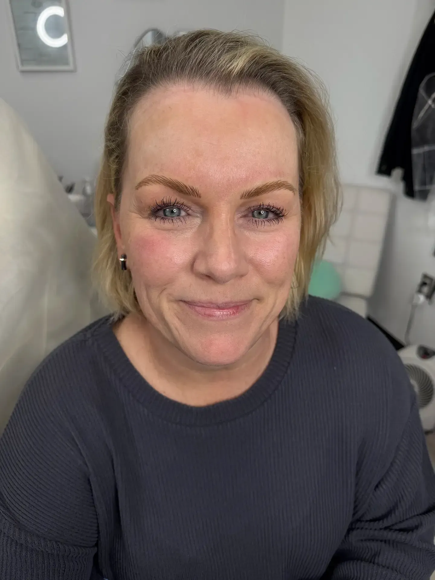 Woman with blonde hair and brown eyebrows smiles at the camera, wearing a gray sweater in a well-lit room.