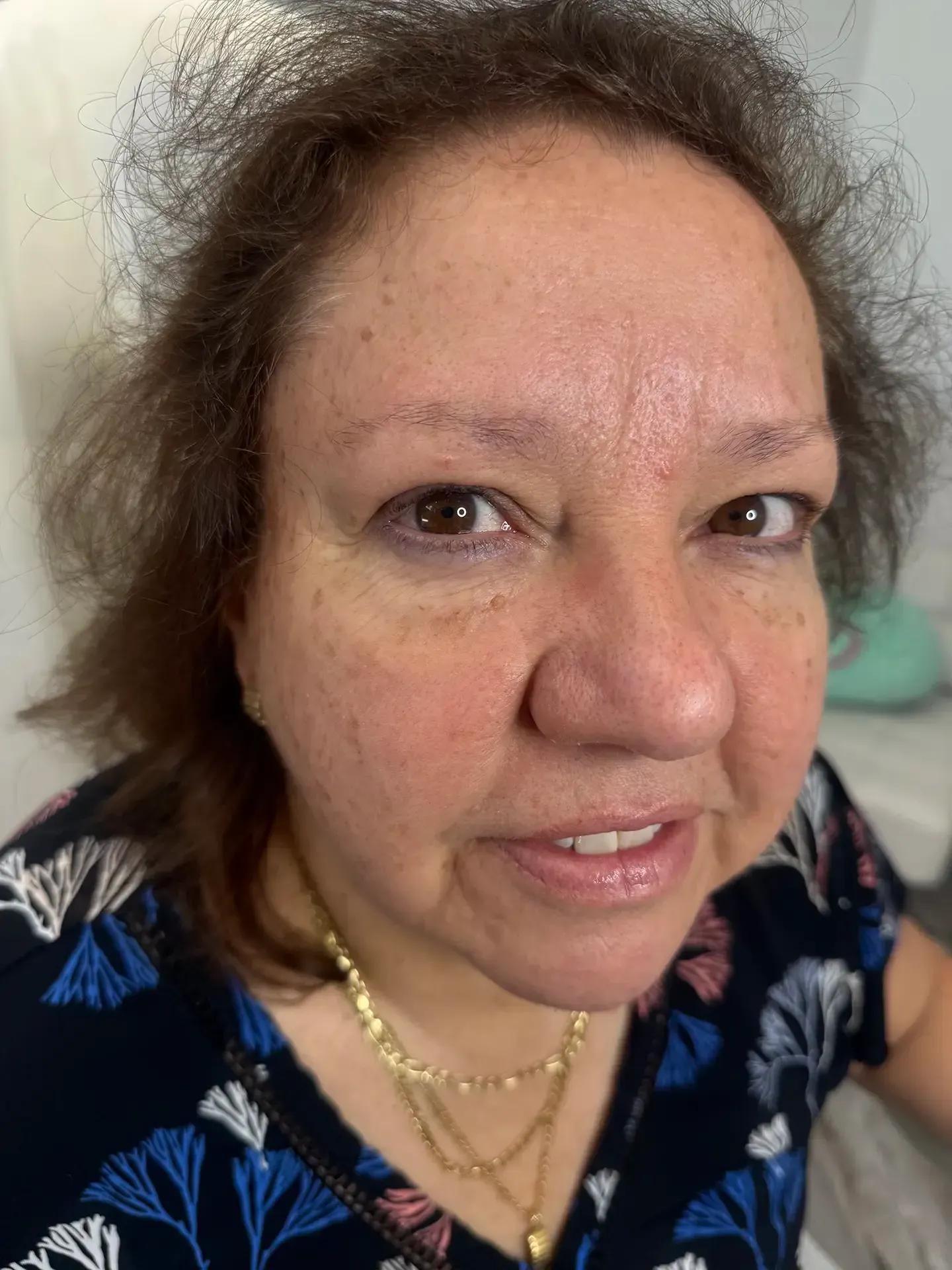 Close-up of a woman with brown eyes, wearing a patterned top and gold necklaces, looking directly at the camera.