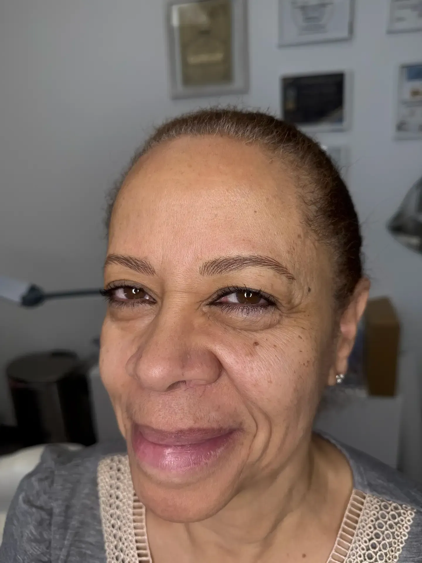 Woman with dark eyebrows and light brown skin smiles, with documents hanging on the white wall behind her.
