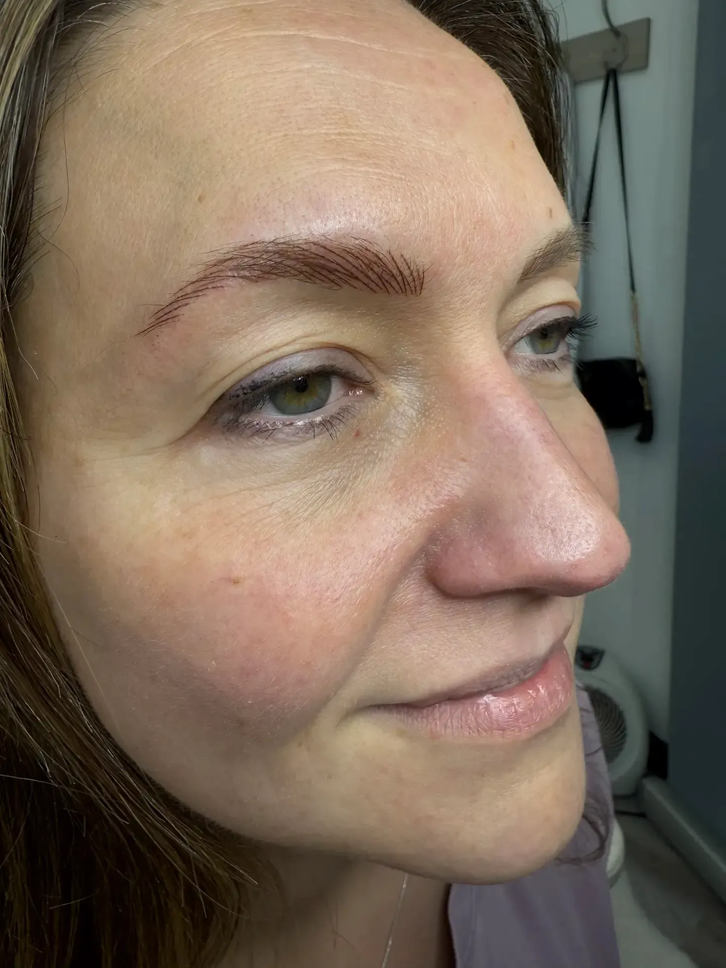Woman with semi-permanent eyebrow makeup, smiling. Close-up view from a side angle, in a beauty salon setting.