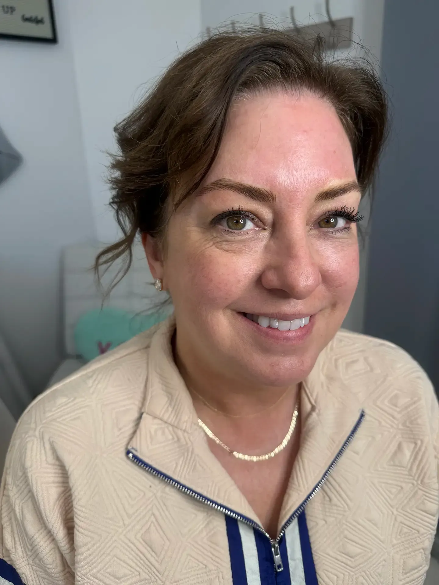 Woman with brown hair smiles, wearing a beige jacket with blue stripes and a gold necklace, in a well-lit room.