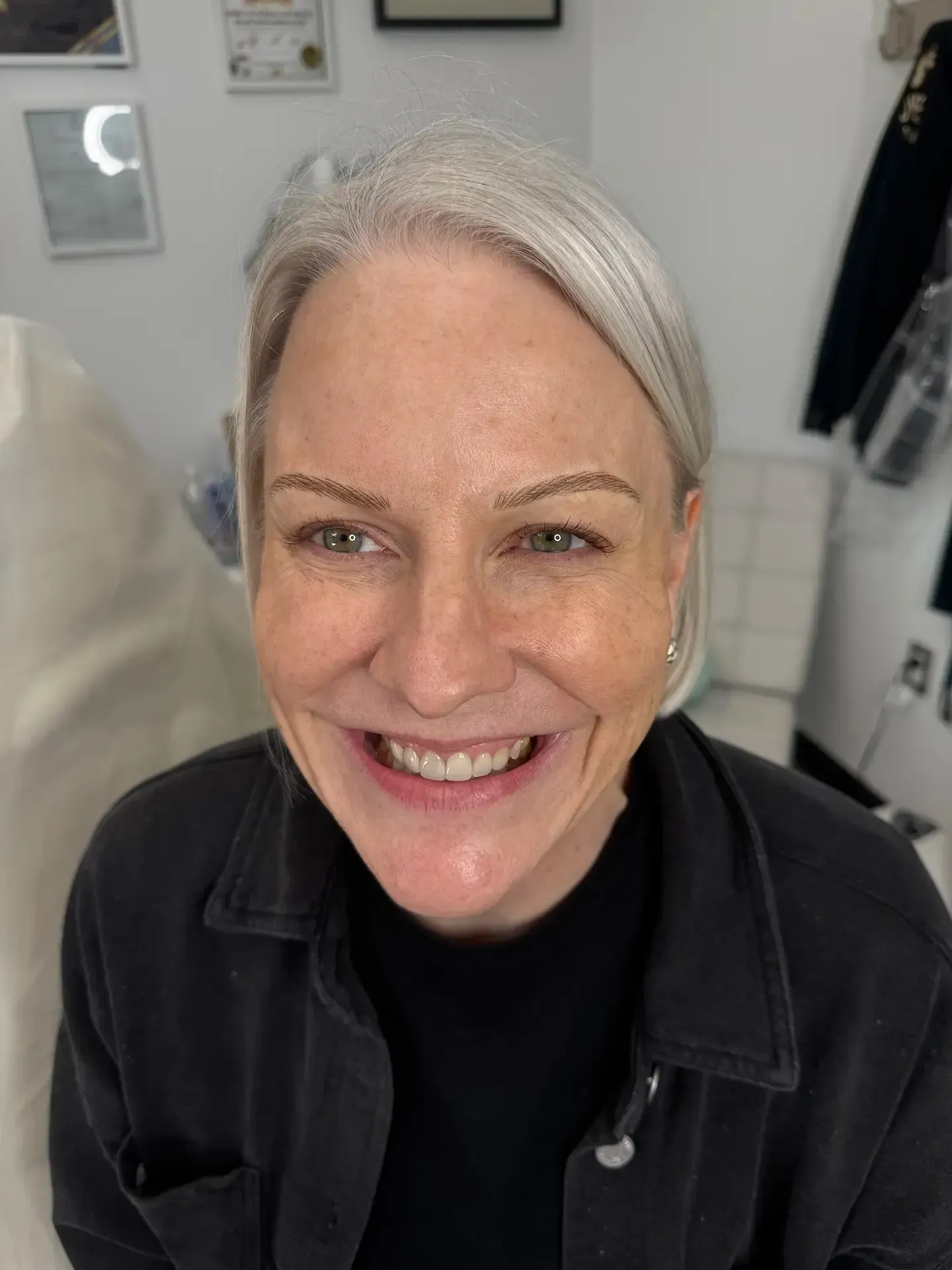 Woman with gray hair smiles at the camera, wearing a black shirt and jacket. She's in a brightly lit room, possibly a clinic.