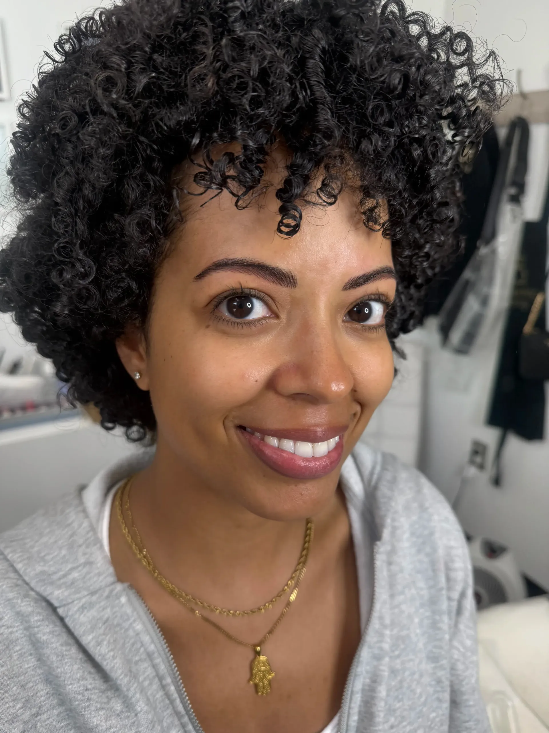Woman with dark, curly hair smiles at the camera, wearing a gray hoodie and gold necklaces. Indoor setting.