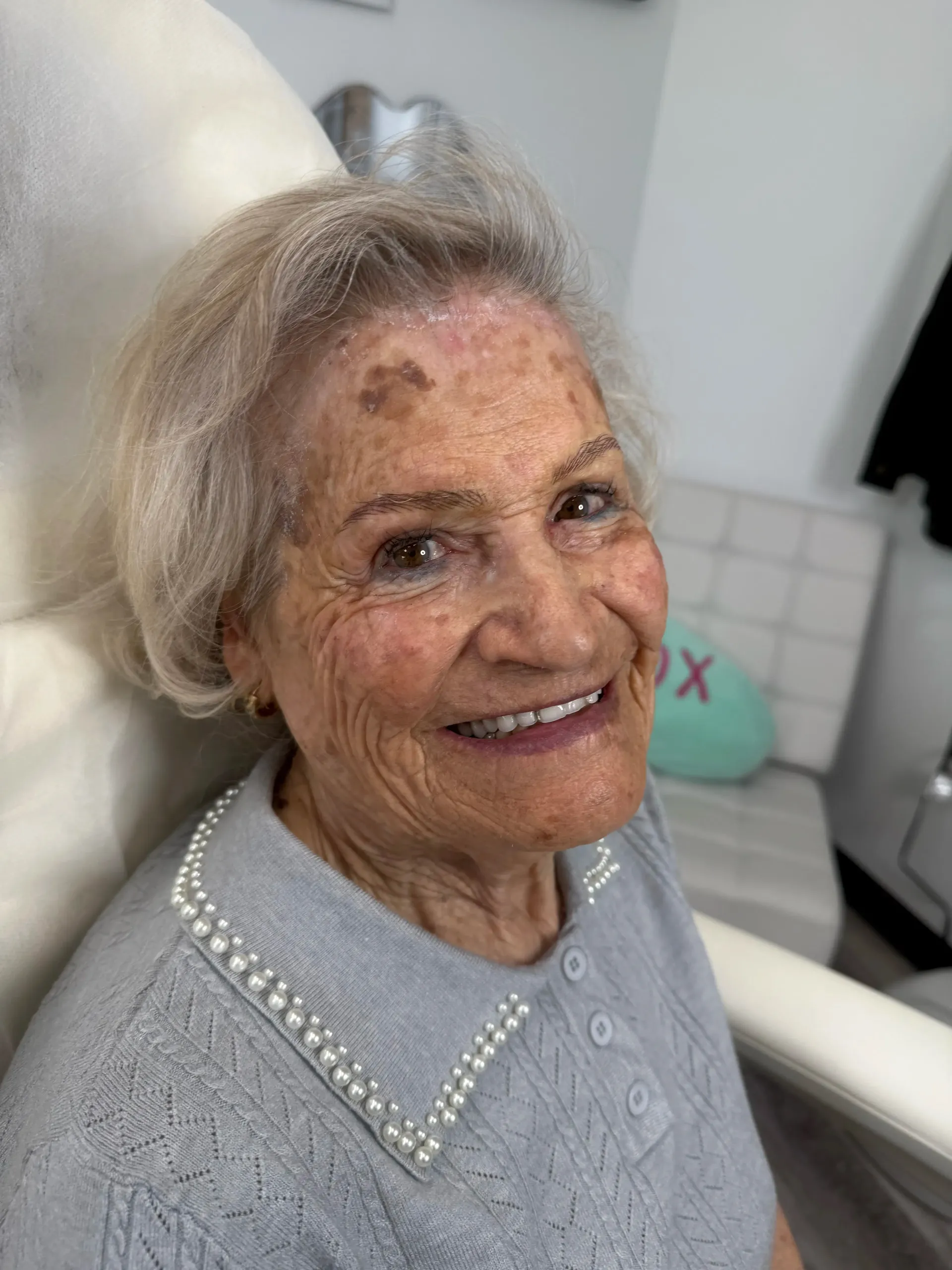 Smiling elderly woman with light hair and a pearl-collared sweater. She has age spots and is sitting inside.