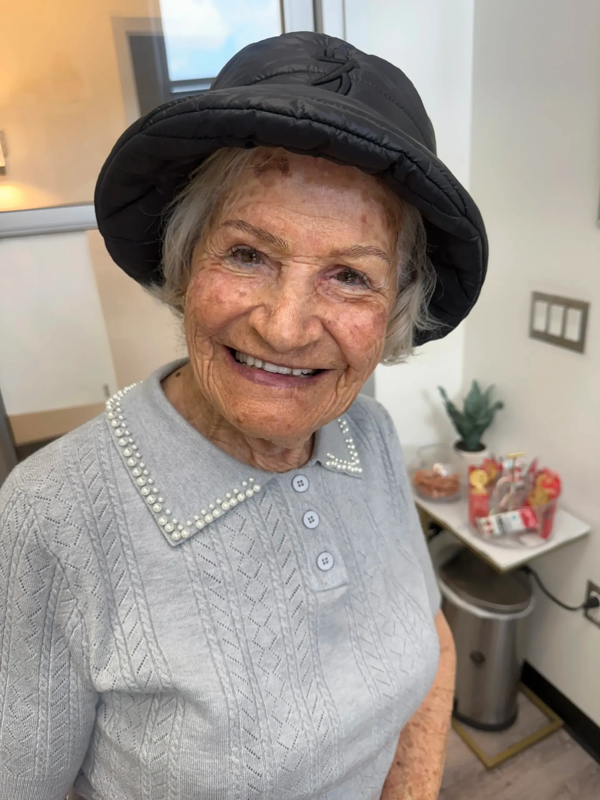 An elderly woman with a smile, wearing a black hat and light gray sweater with pearl-like trim, indoors.