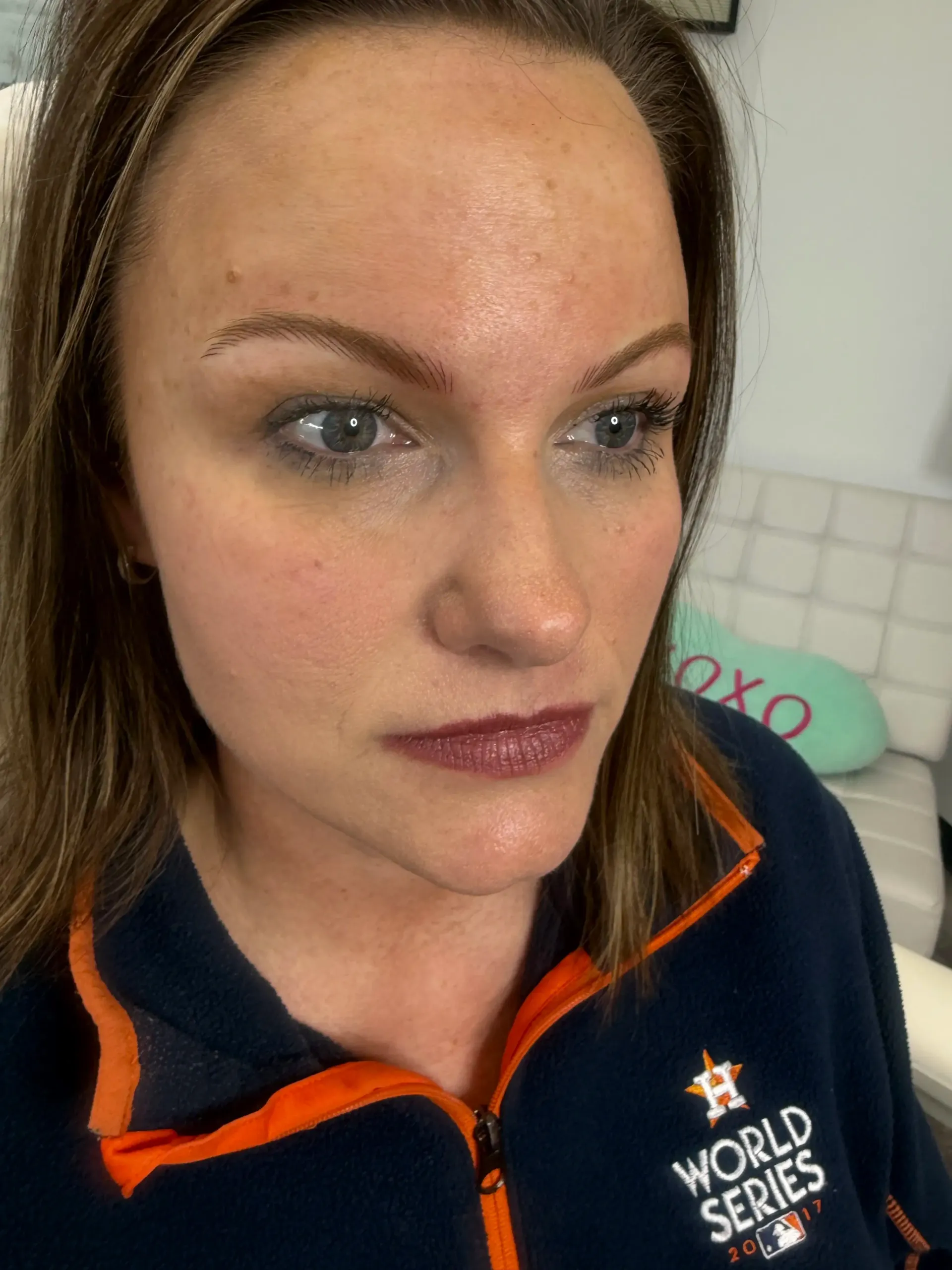 Woman with light makeup and brown eyebrows, wearing a navy Houston Astros World Series sweatshirt, looking off to the side.