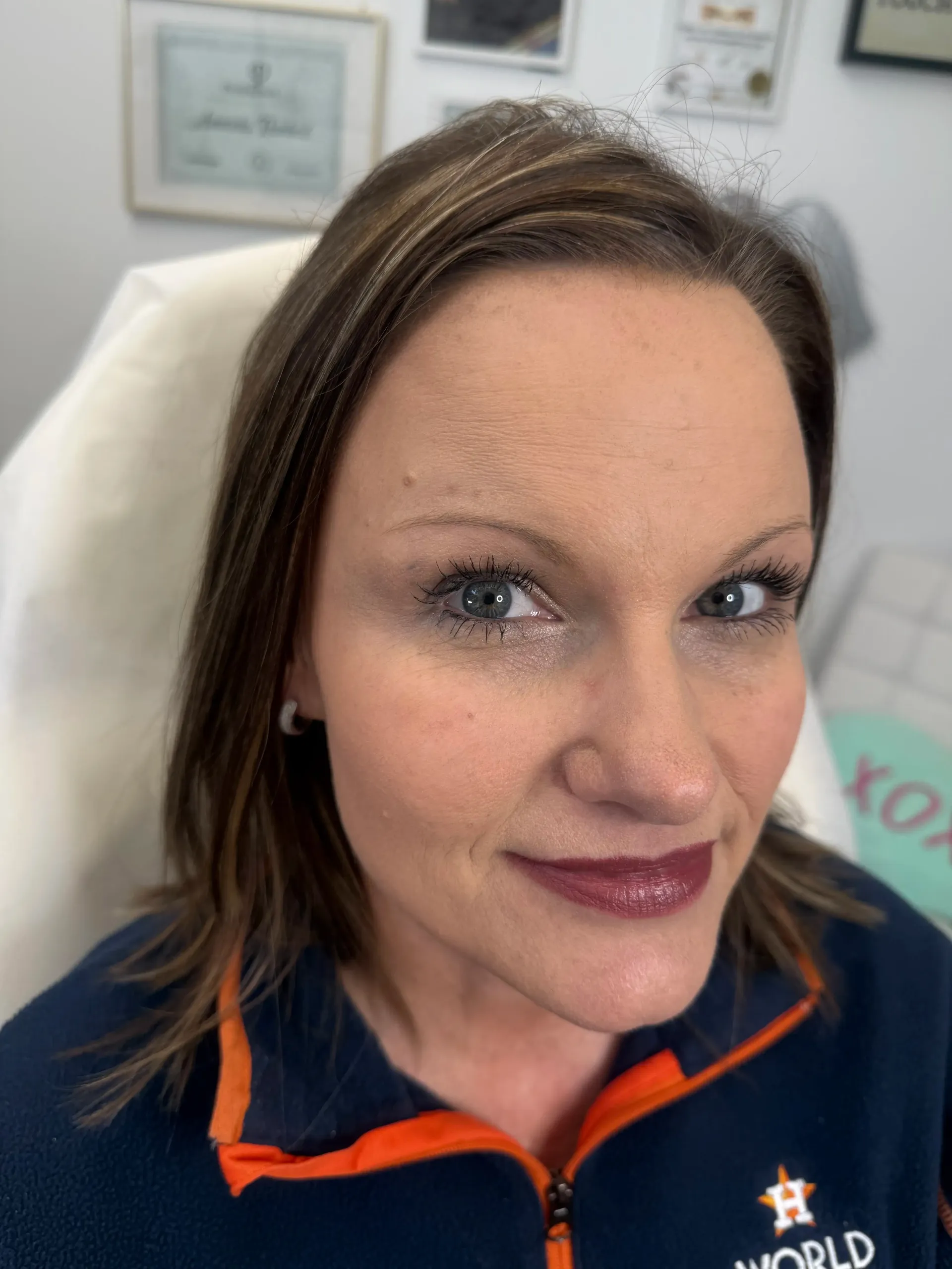 Woman with dark lipstick and glitter makeup, seated in a chair. She wears a navy and orange Houston Astros jacket.