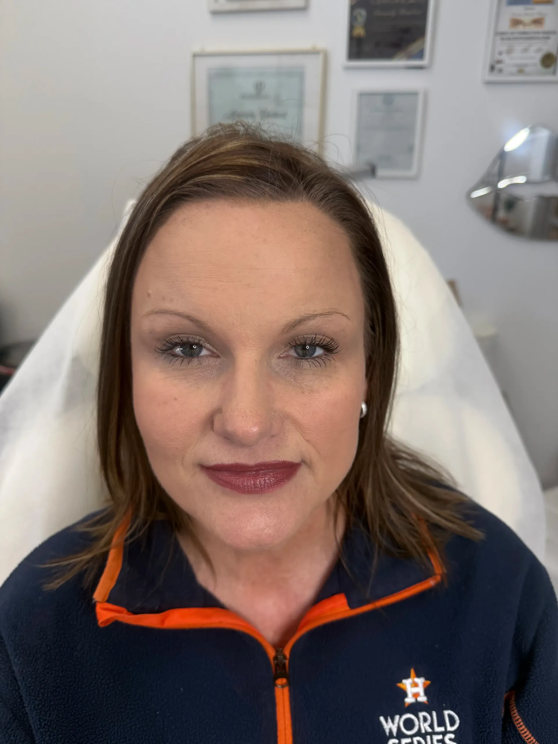 Woman with makeup sitting in a medical chair. She has dark hair, a neutral expression, and a dark blue jacket with an orange logo.