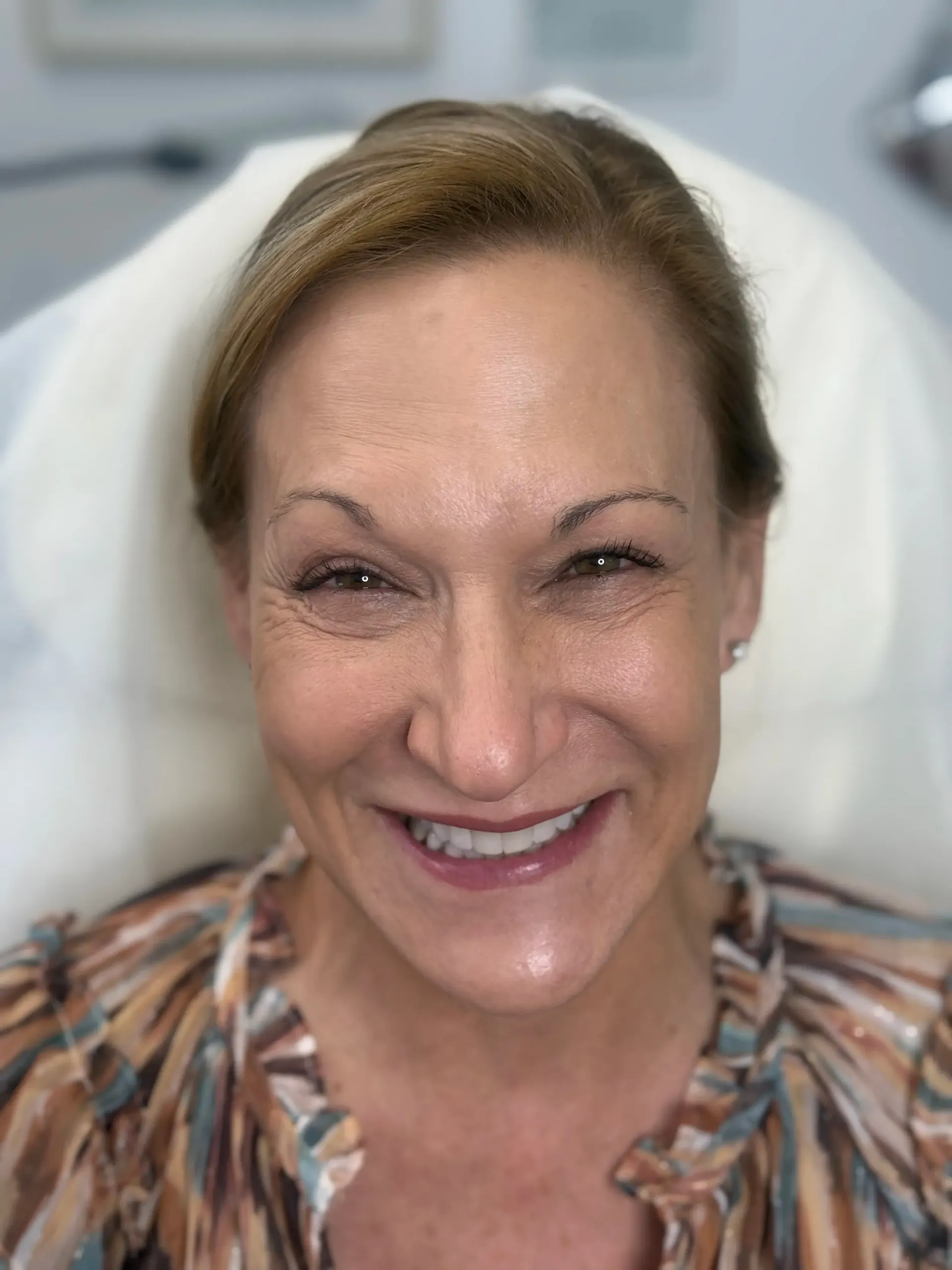 Smiling woman with brown hair and a patterned shirt; she is in a medical setting.