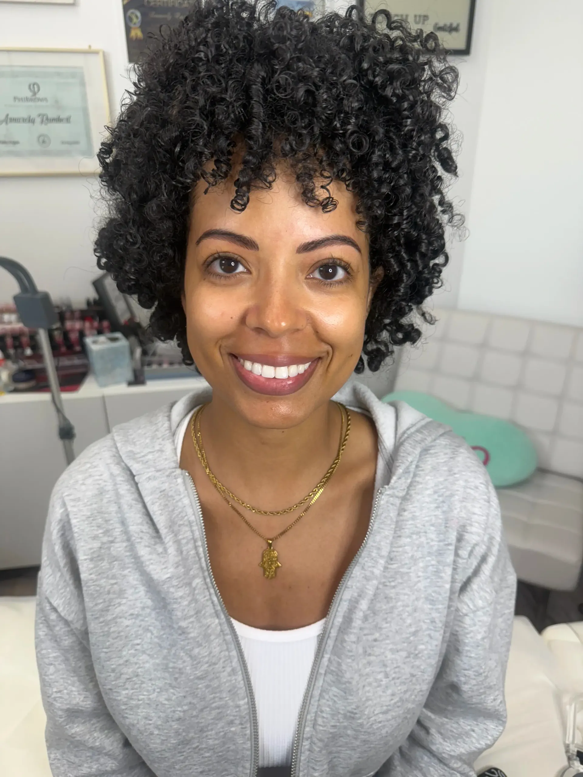 A woman with curly dark hair, wearing a gray hoodie, smiling at the camera. She has gold necklaces and is in a brightly lit room.