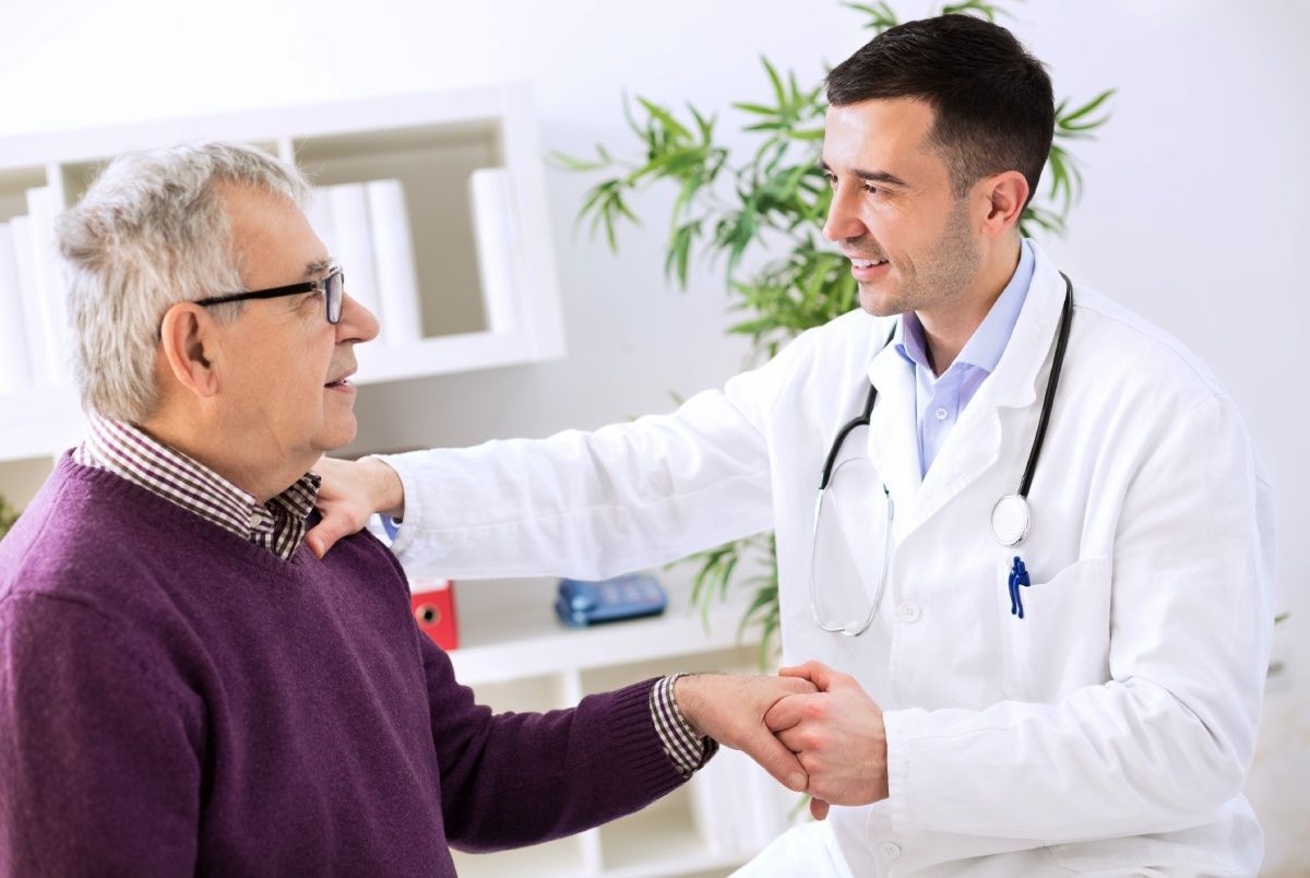 Doctor in white coat smiles, comforts older patient in purple sweater; touching hands, office setting.