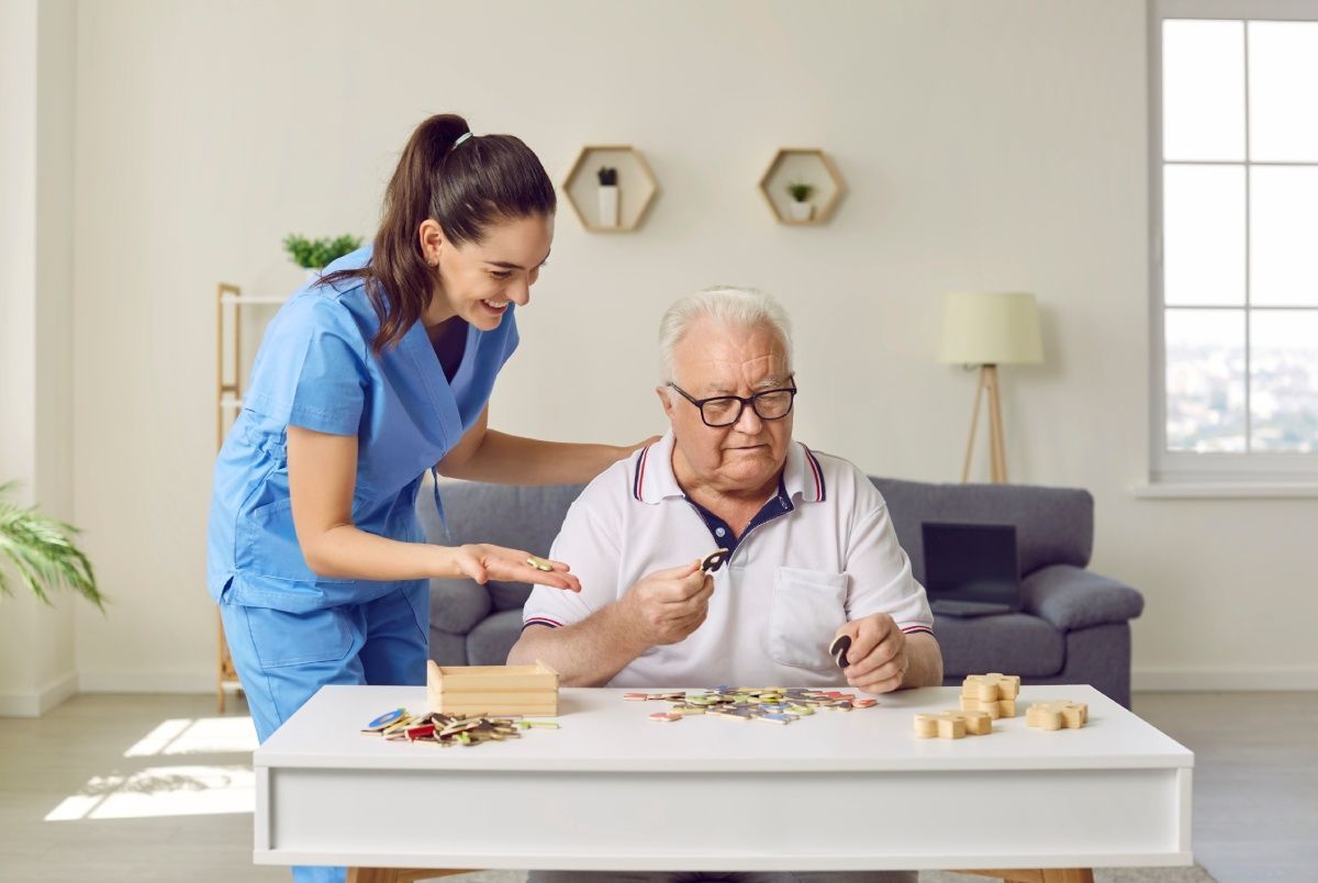 Caregiver assisting senior with a puzzle. They are in a living room; the caregiver is smiling.