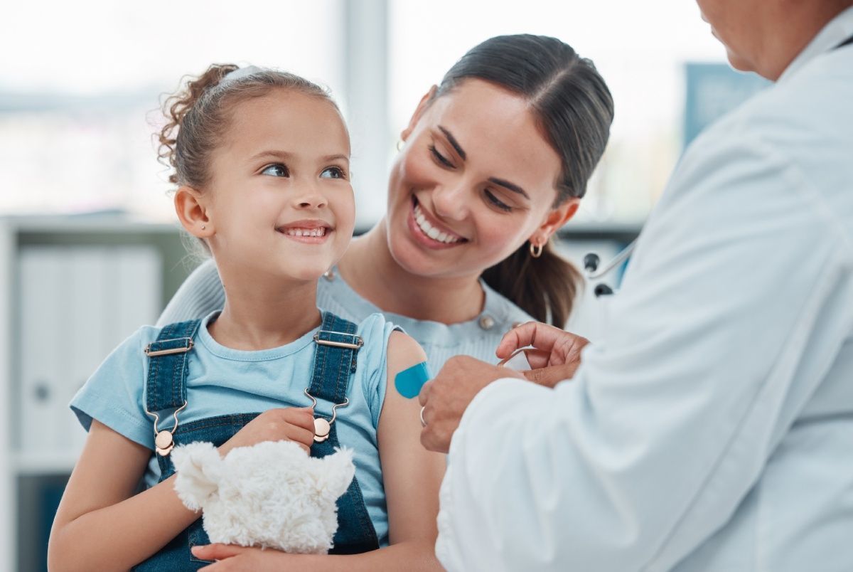 A child smiles as a doctor applies a bandage after a shot, with a smiling parent present.