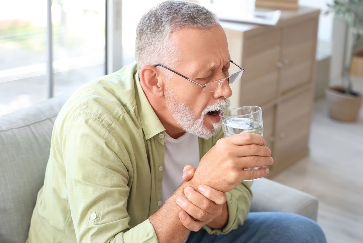 Man coughing, holding glass of water, indoors, seated on couch.