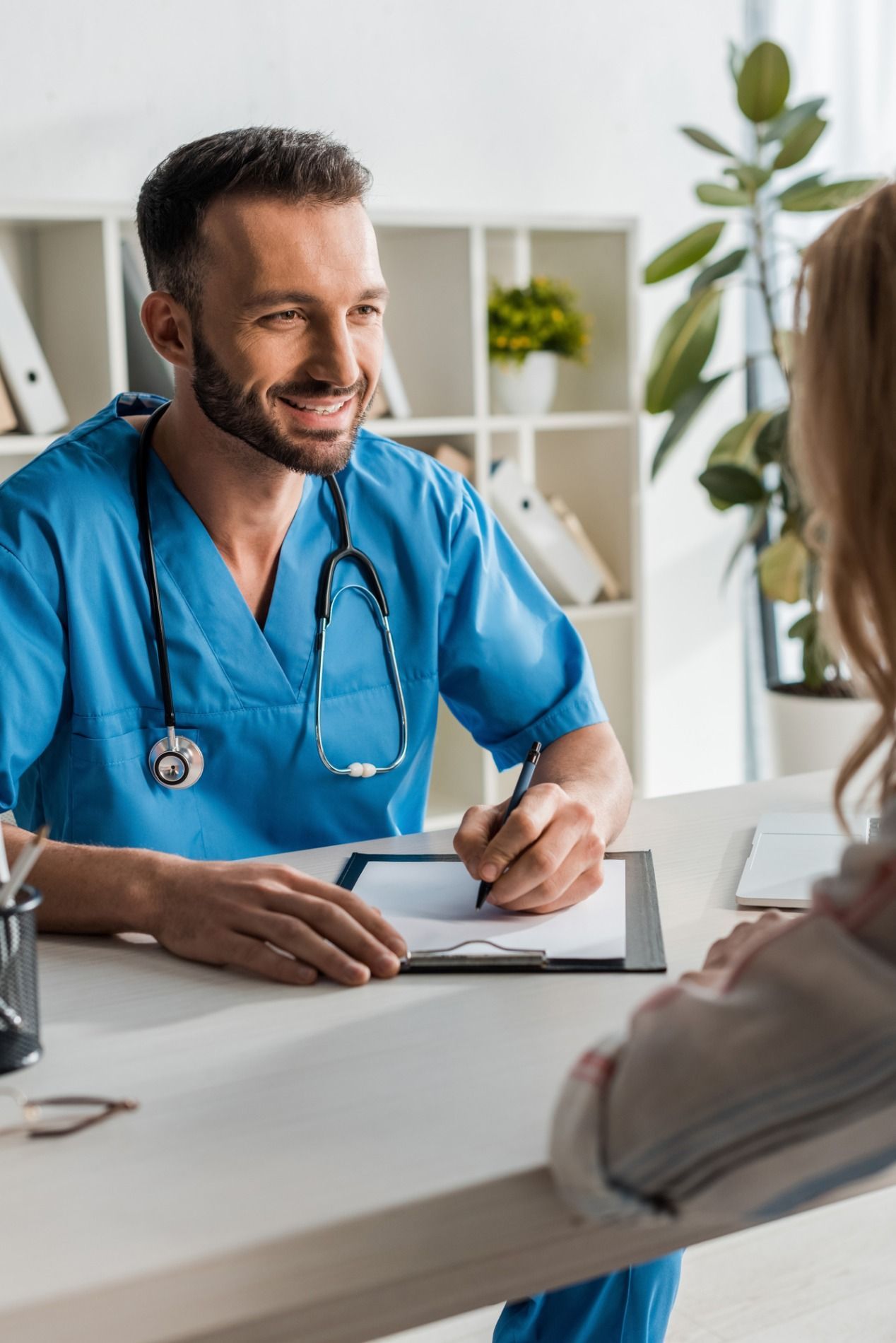 Doctor in blue scrubs smiles while taking notes during a consultation with a patient.