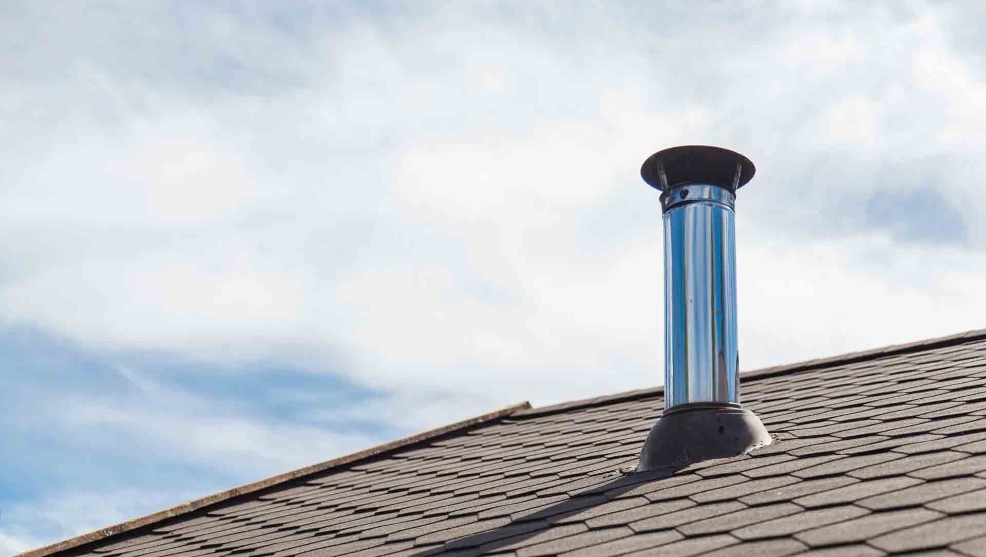 Un tuyau de cheminée en acier inoxydable dépasse d'un toit de bardeaux bruns sur un ciel bleu nuageux.