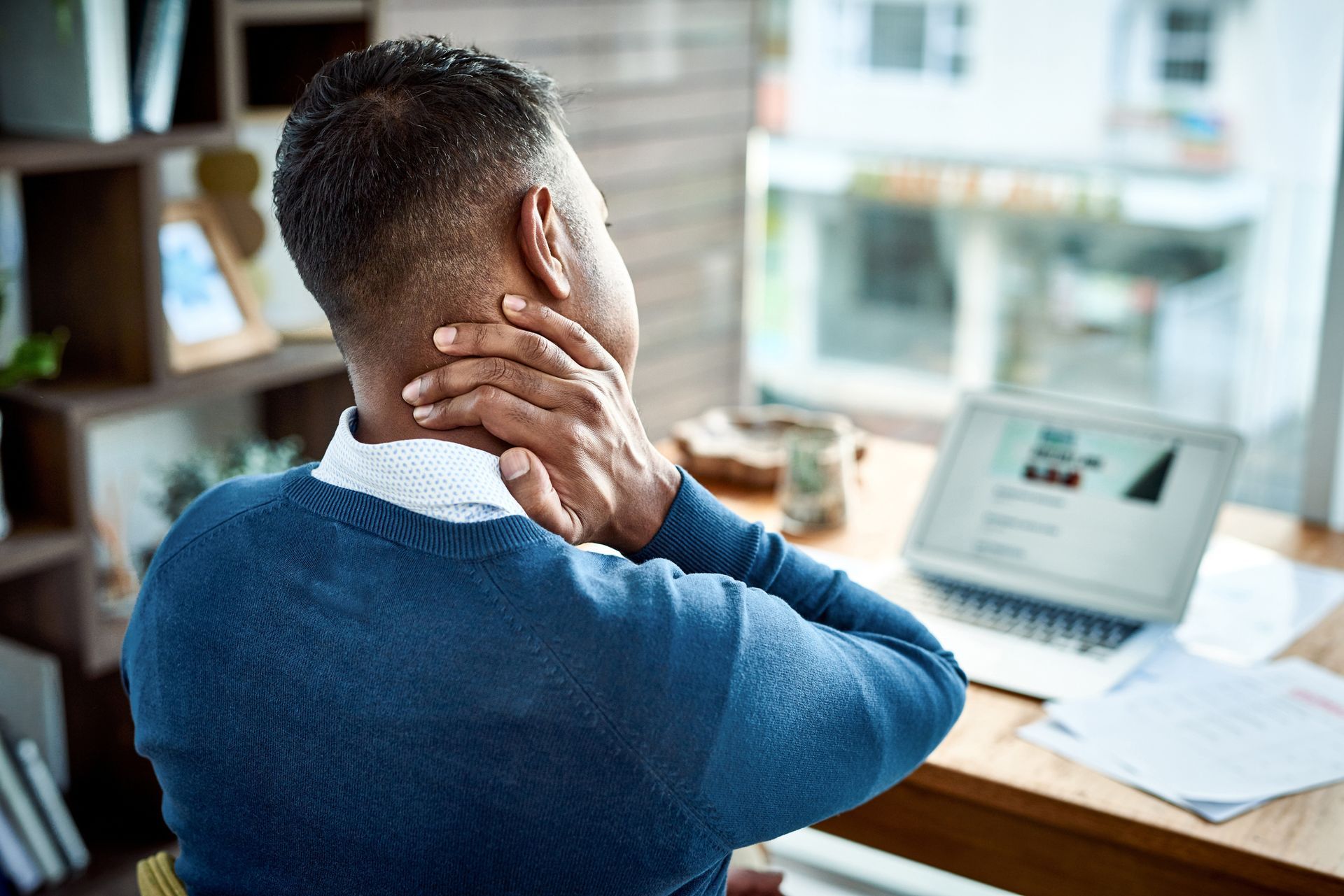 Man sitting at a desk, clutching his neck, likely experiencing pain. Laptop and papers nearby.