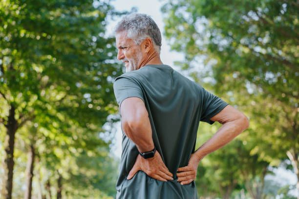 Older man outdoors, clutching his lower back in pain. Green shirt, trees in background.