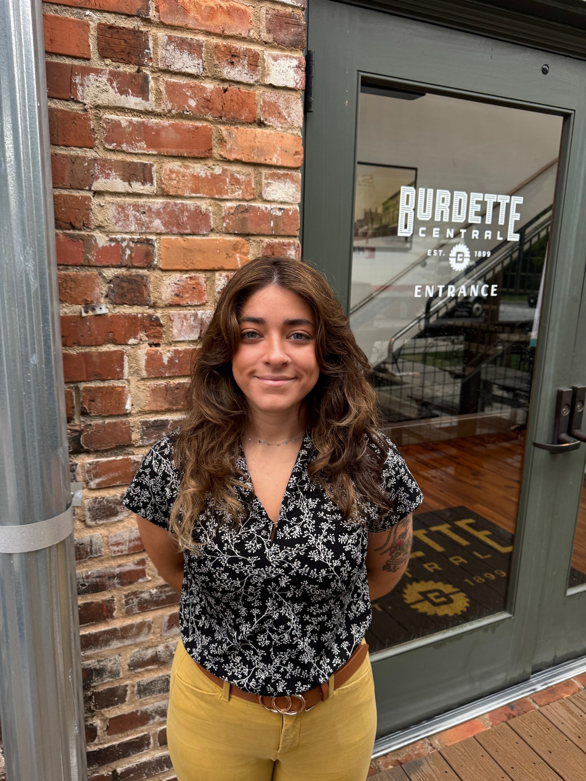 Woman with brown curly hair, in front of a brick wall and glass door of a business, smiling.