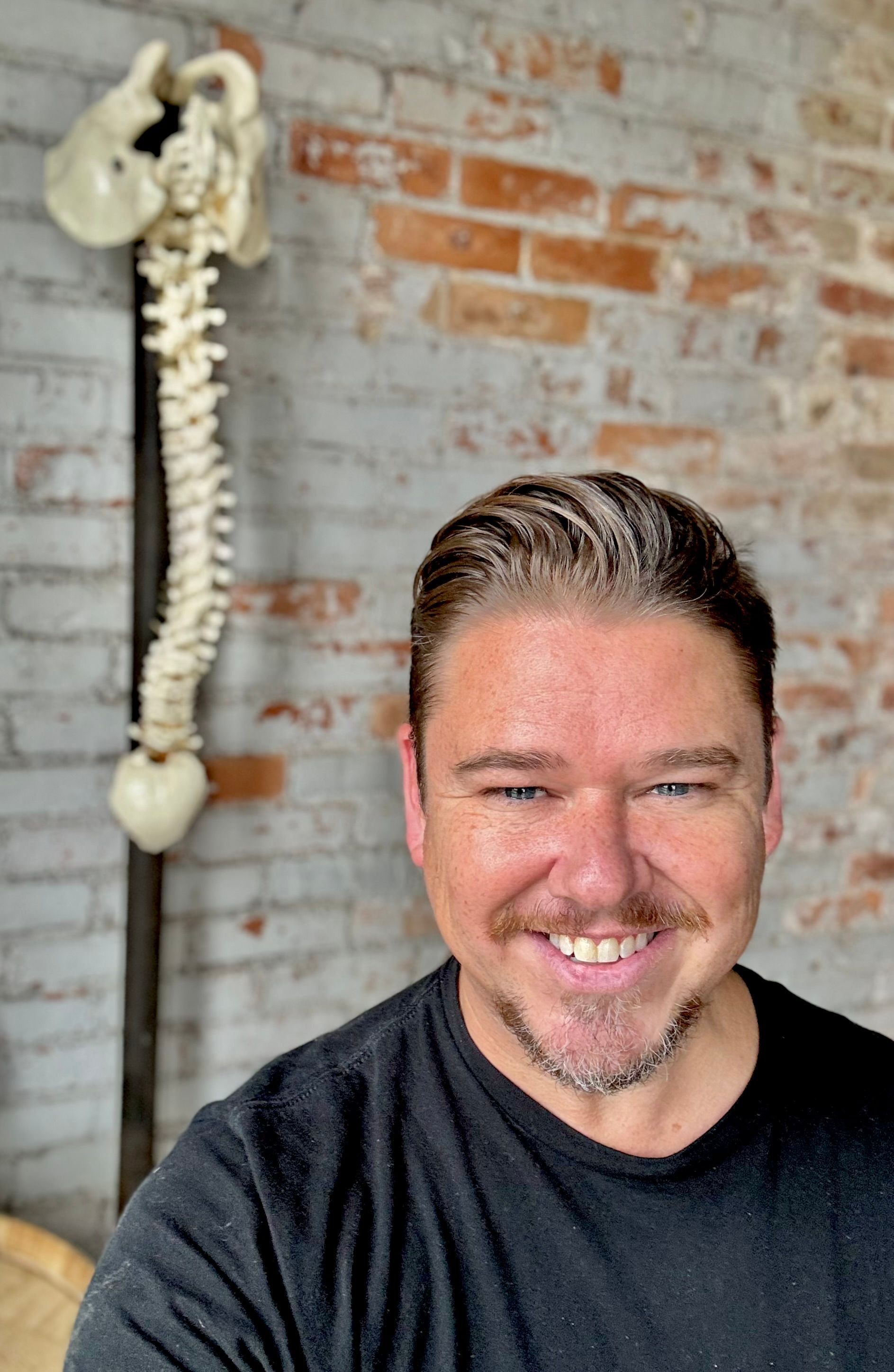Man smiles at the camera, with a spine model hanging on a brick wall in the background.
