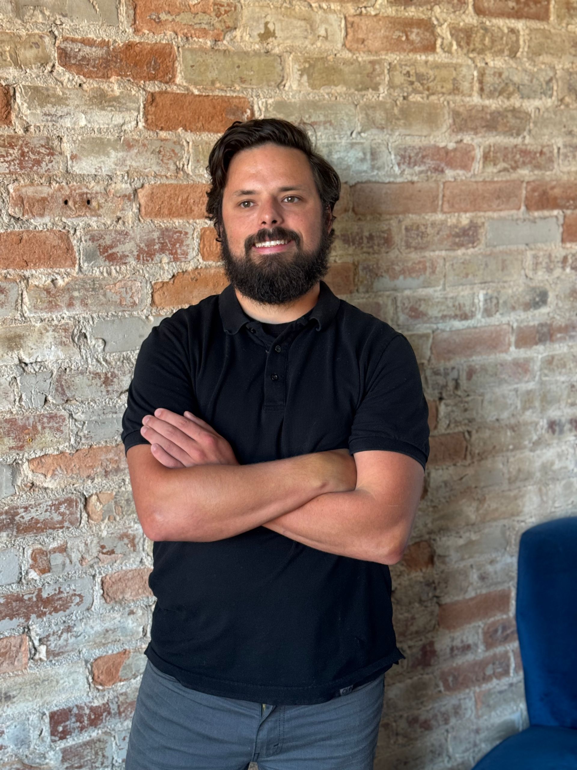 Man with beard, arms crossed, standing in front of a brick wall. Wearing black shirt and gray pants.