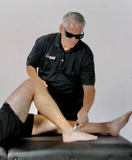 Man in sunglasses using a device on a patient's ankle. Both are on a black table in a clinic setting.