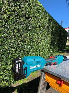 A makita pole saw is sitting on a table in front of a hedge.