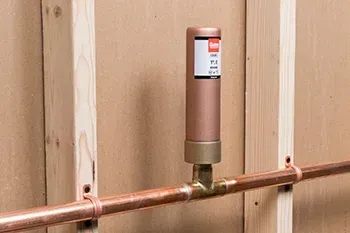A copper water hammer arrestor installed on a horizontal copper pipe mounted to wooden wall studs.