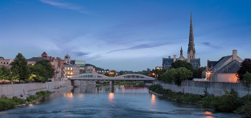 A city skyline at dusk, with a river in the foreground and a bridge, church spire, and buildings.