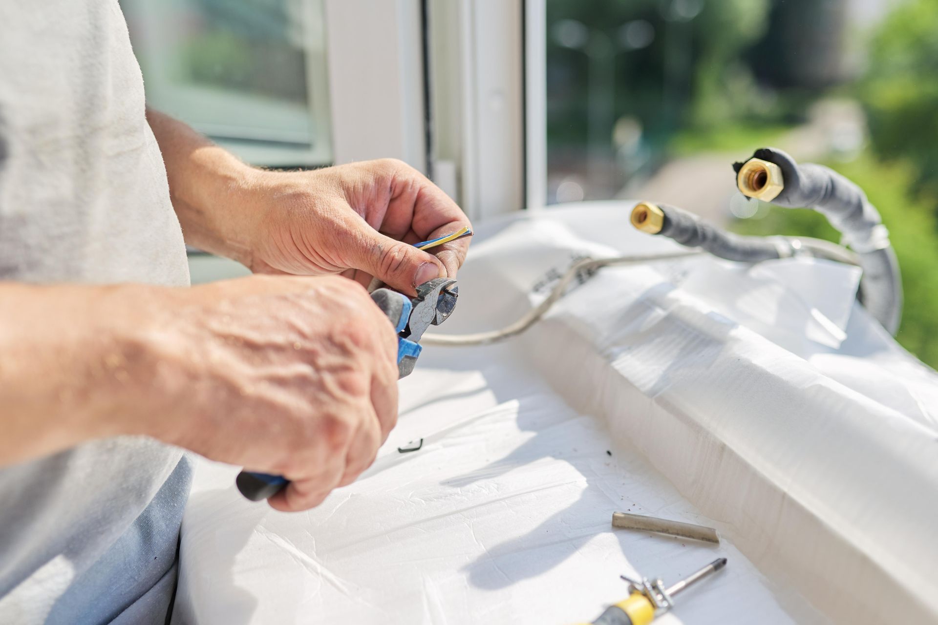 Person using pliers to cut a wire near a window with AC unit components visible.