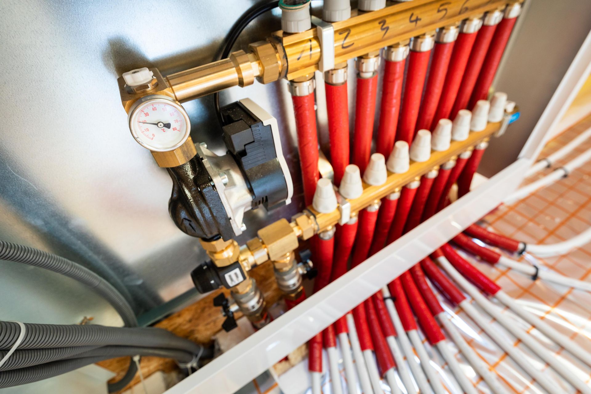Close-up of radiant floor heating manifold with red and white pipes connected. A gauge and pump are visible.