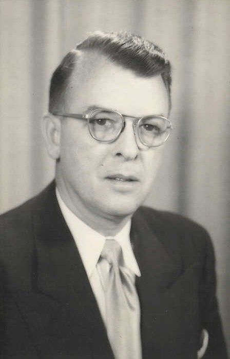 A formal black-and-white portrait of a man wearing glasses, a suit, and a tie, posed against a curtained backdrop.