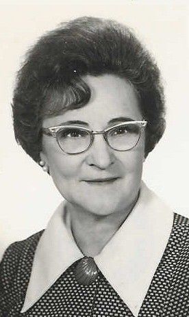 A black-and-white portrait of a person with styled hair wearing glasses, a collared dress, and a circular brooch.