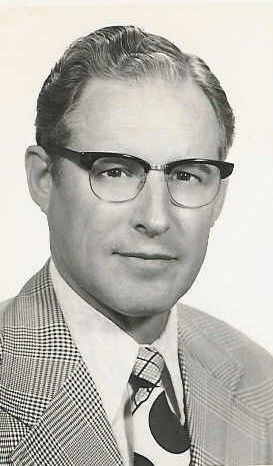 A studio portrait of a person wearing glasses, a patterned blazer, and a polka-dot tie against a light background.