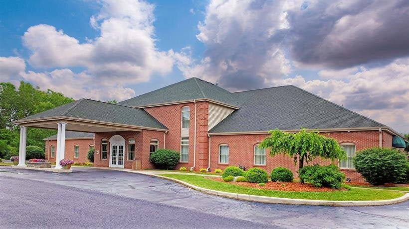 A single-story red brick building with a dark shingled roof, a covered entrance, and green shrubs under a cloudy sky.
