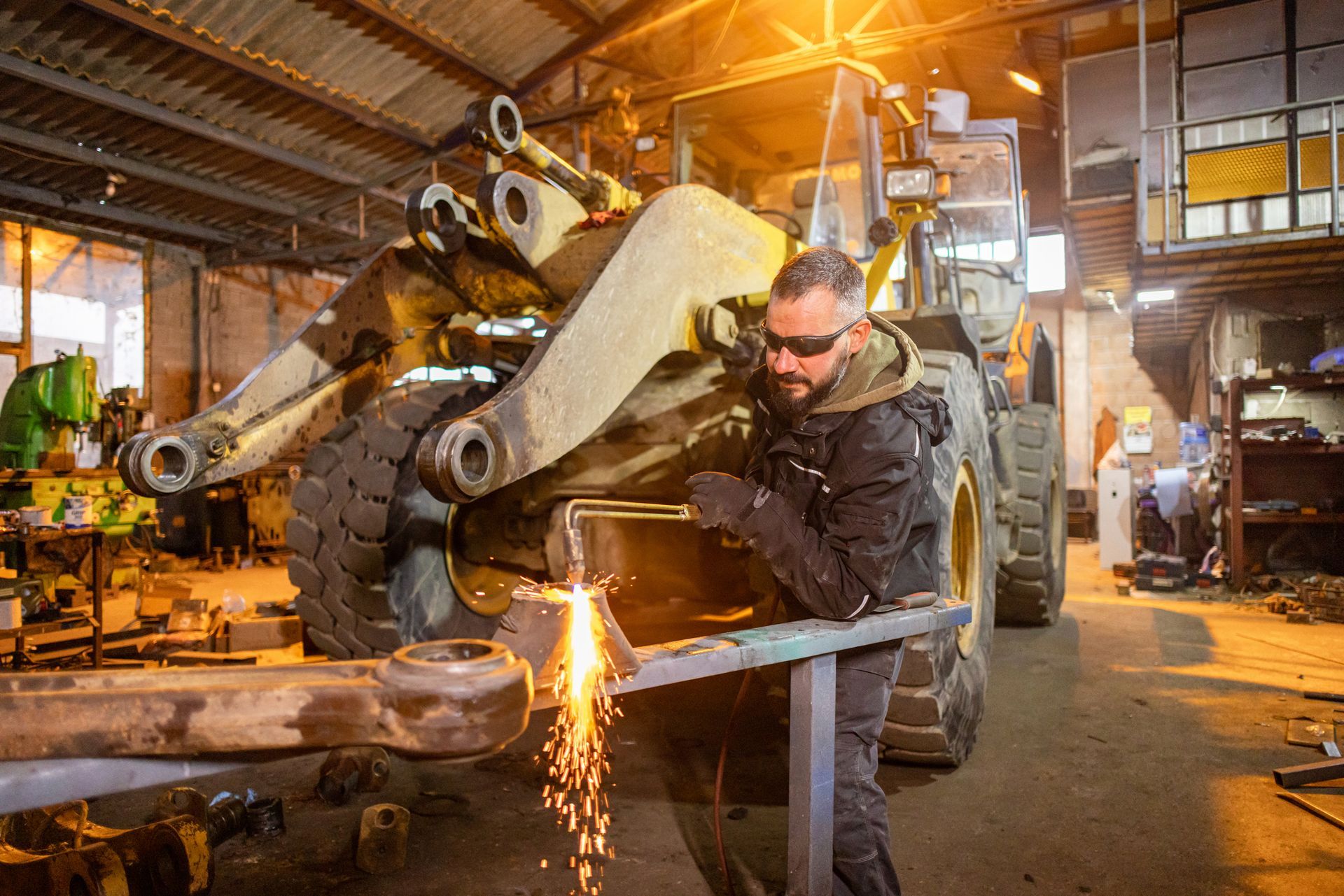 Mechanic welding heavy machinery in workshop with flying sparks and industrial equipment background.