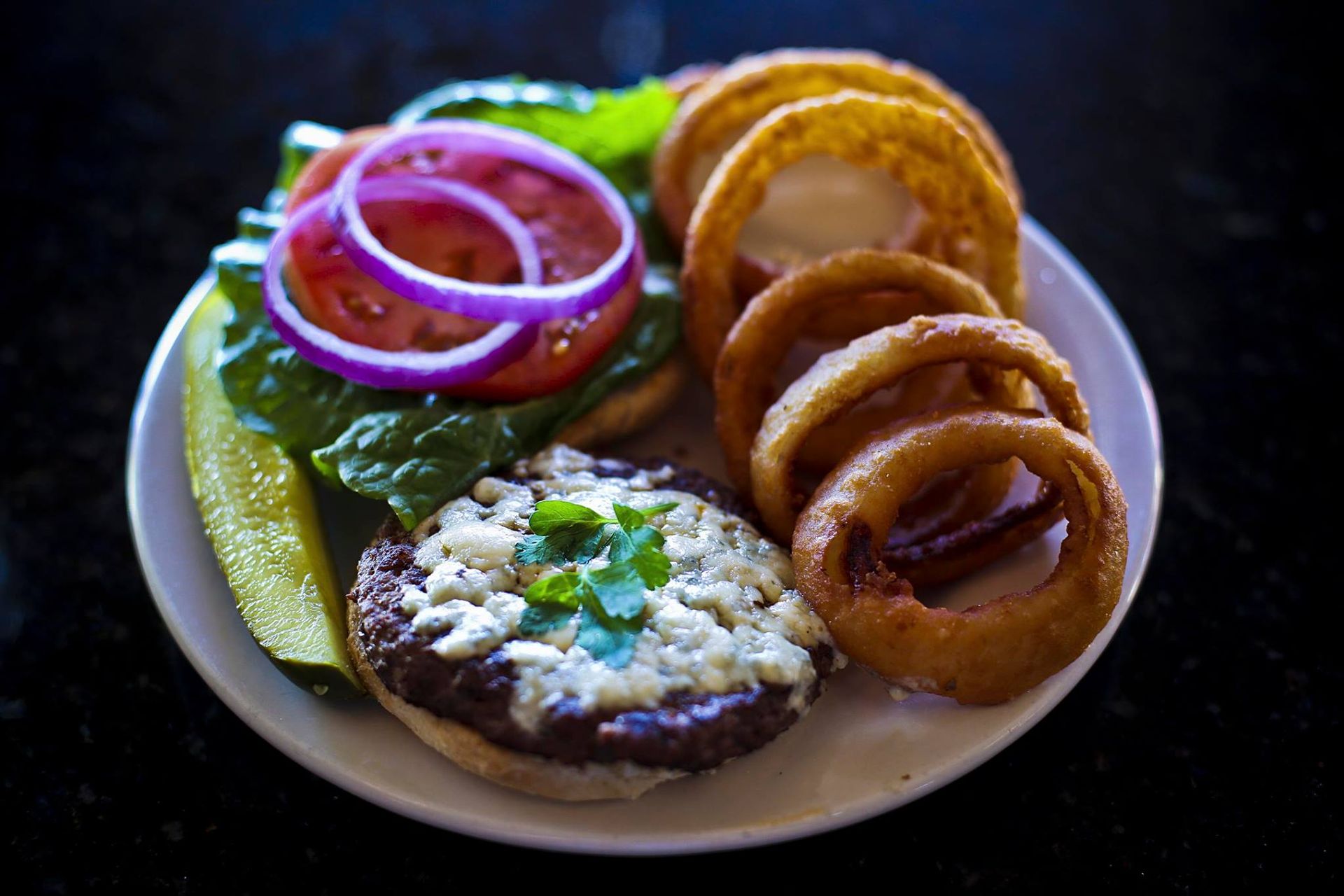 Burger topped with cheese, tomato, and onion, served with onion rings and a pickle on a white plate.