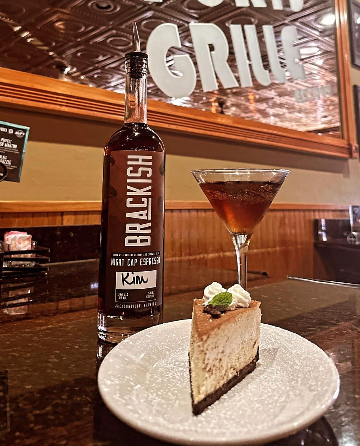 A bottle of Brackish spirits next to a cocktail and a slice of cheesecake on a bar. The setting appears to be a restaurant.