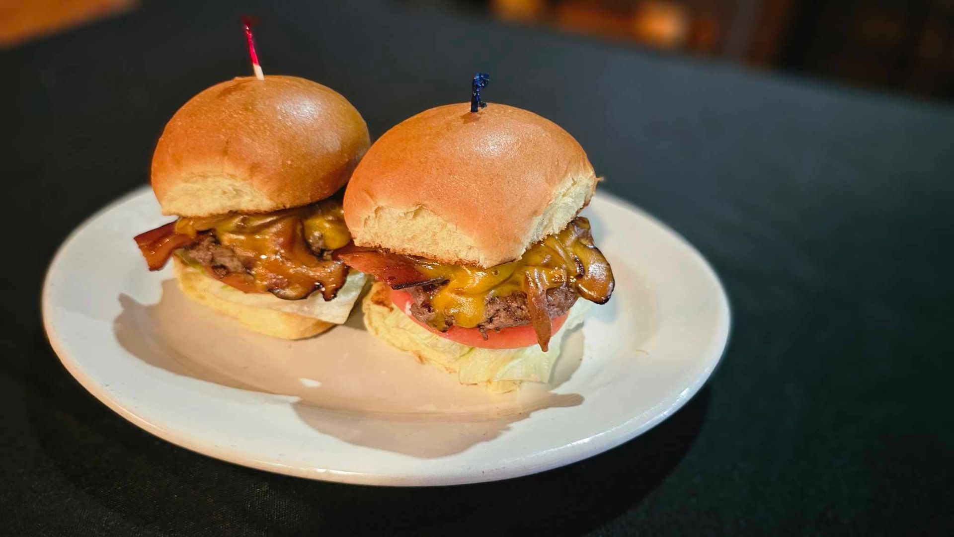 Two mini burgers on a white plate, with bacon, cheese, and a toothpick. Set on a black surface.