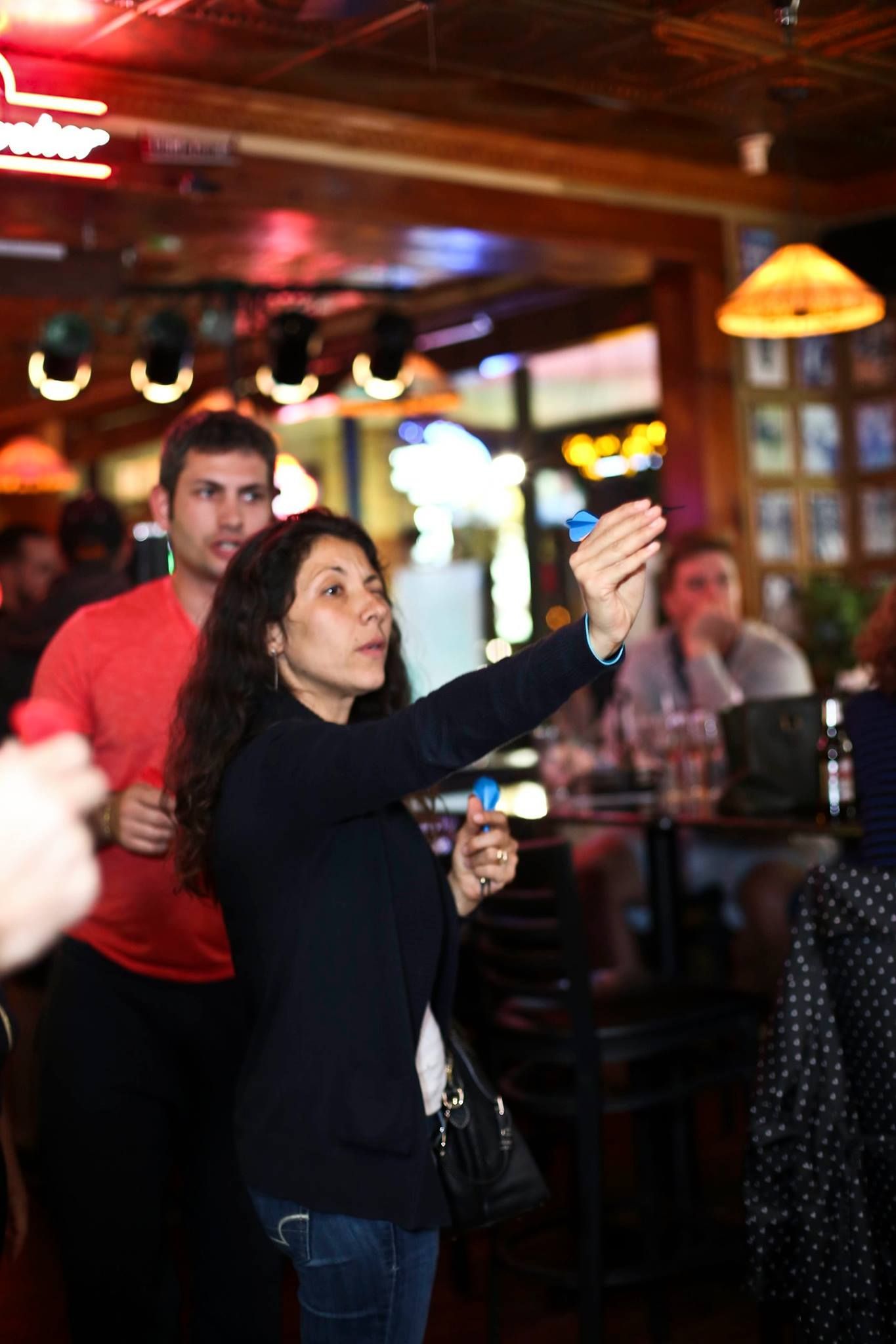 Woman in a dark blue sweater raises her arm in a bar, with a man in a red shirt behind her.
