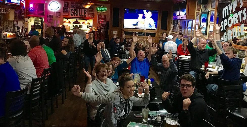 People cheering in a crowded sports bar with multiple TVs showing a game. Many have their arms raised in excitement.