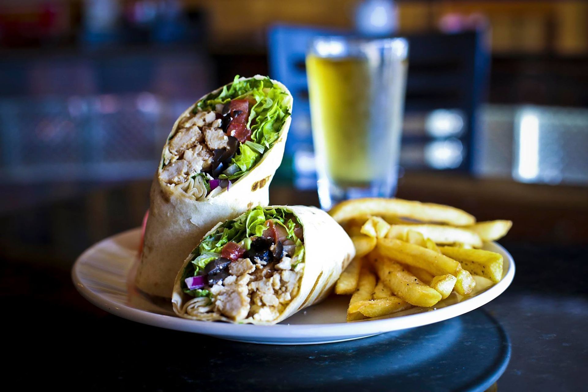 Chicken wrap cut in half with fries and a glass of beer on a white plate.