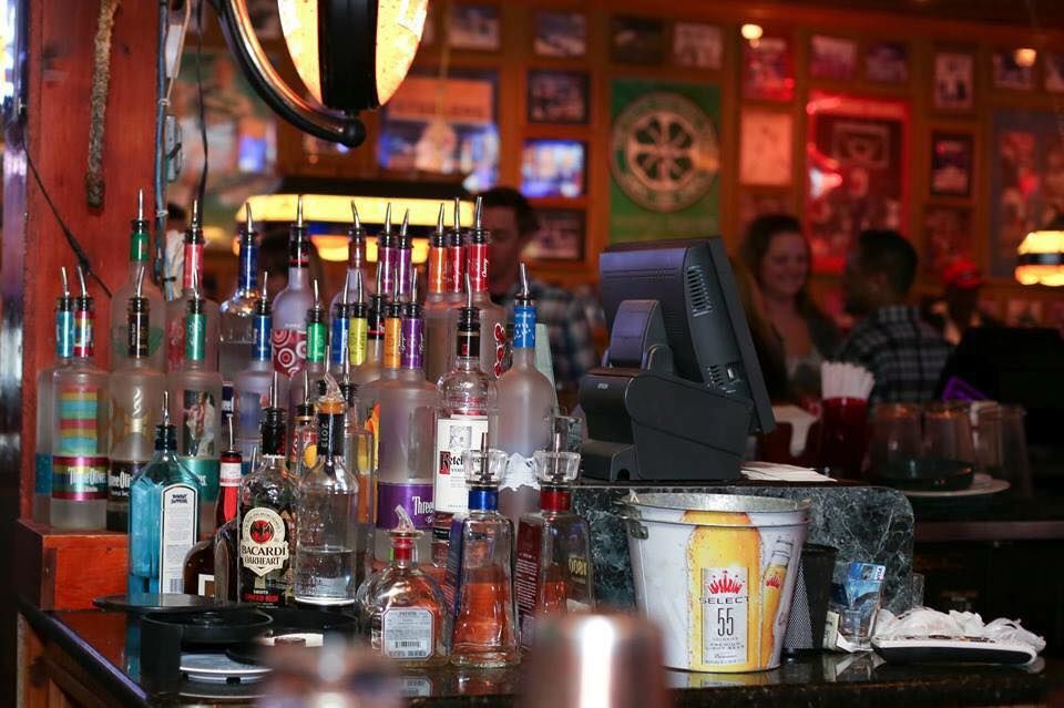 Bar scene: Bottles of liquor line a counter. Behind, blurred people chat near a cash register and TV screens.