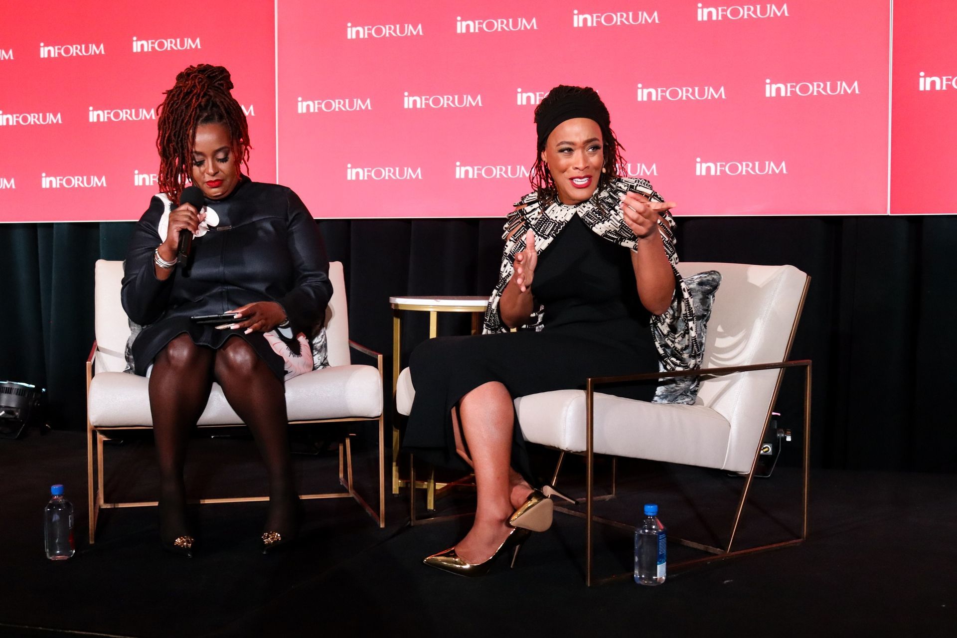 Two women are sitting in chairs on a stage talking to each other.