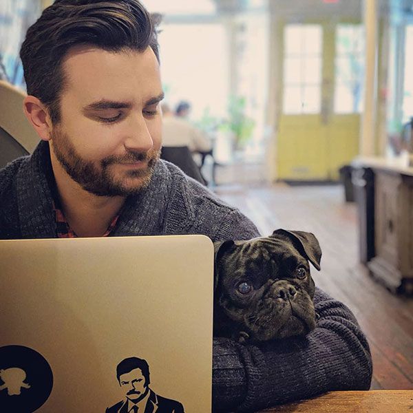 A man is sitting at a table with a laptop and a dog on his lap.