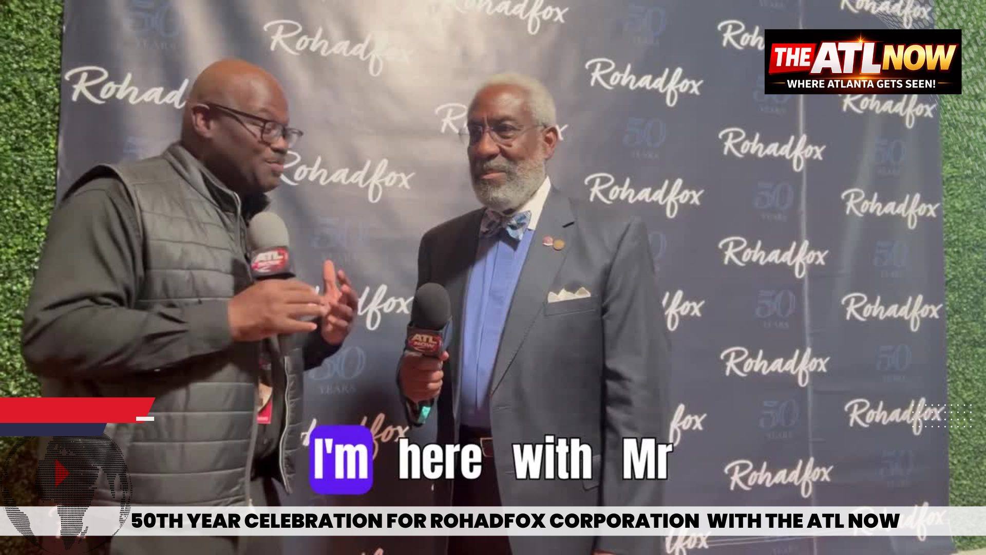 Two men talking at a Rohabox event backdrop, one holding a drink, with event banner text visible.