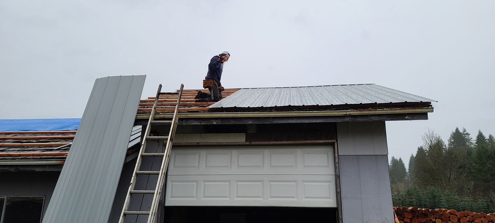 Person on a roof replacing metal panels. A ladder leans against the building. Overcast sky.