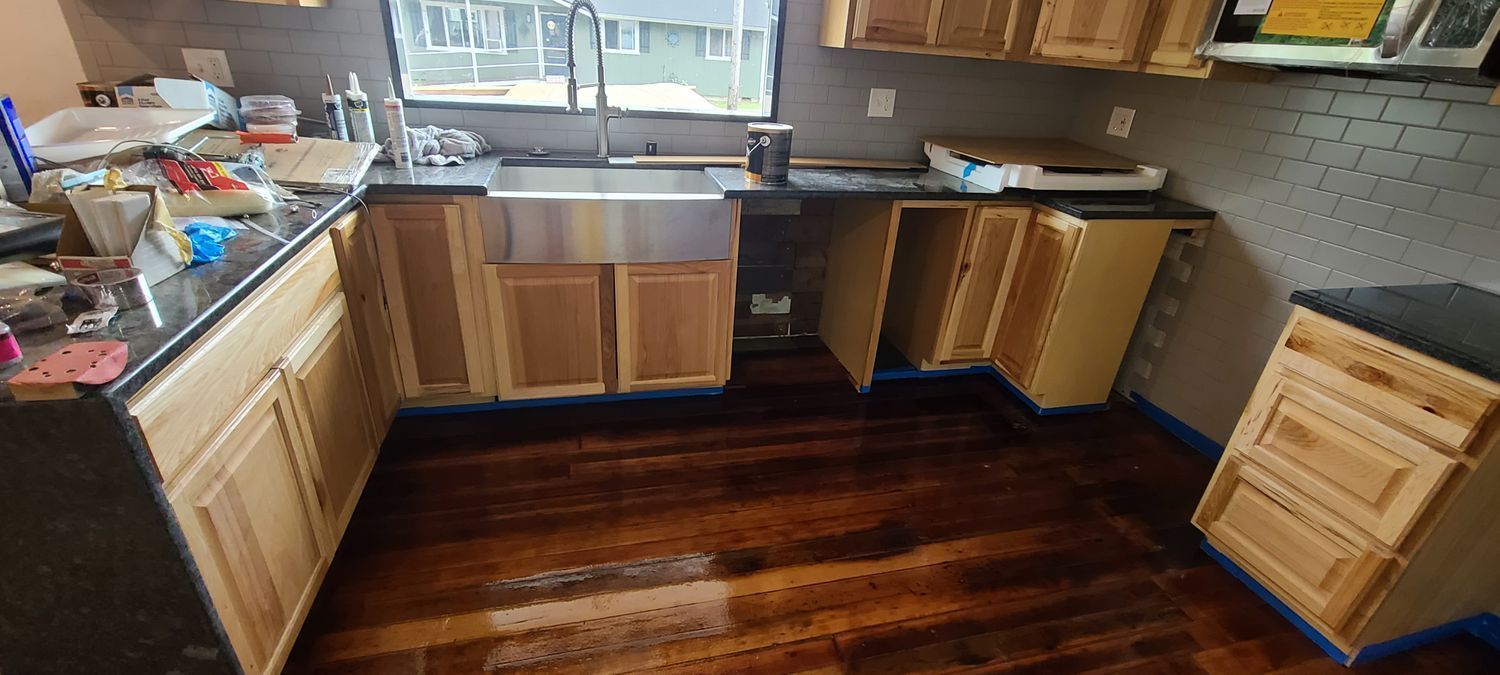 Kitchen undergoing renovation with exposed cabinets, dark countertops, and wood flooring.