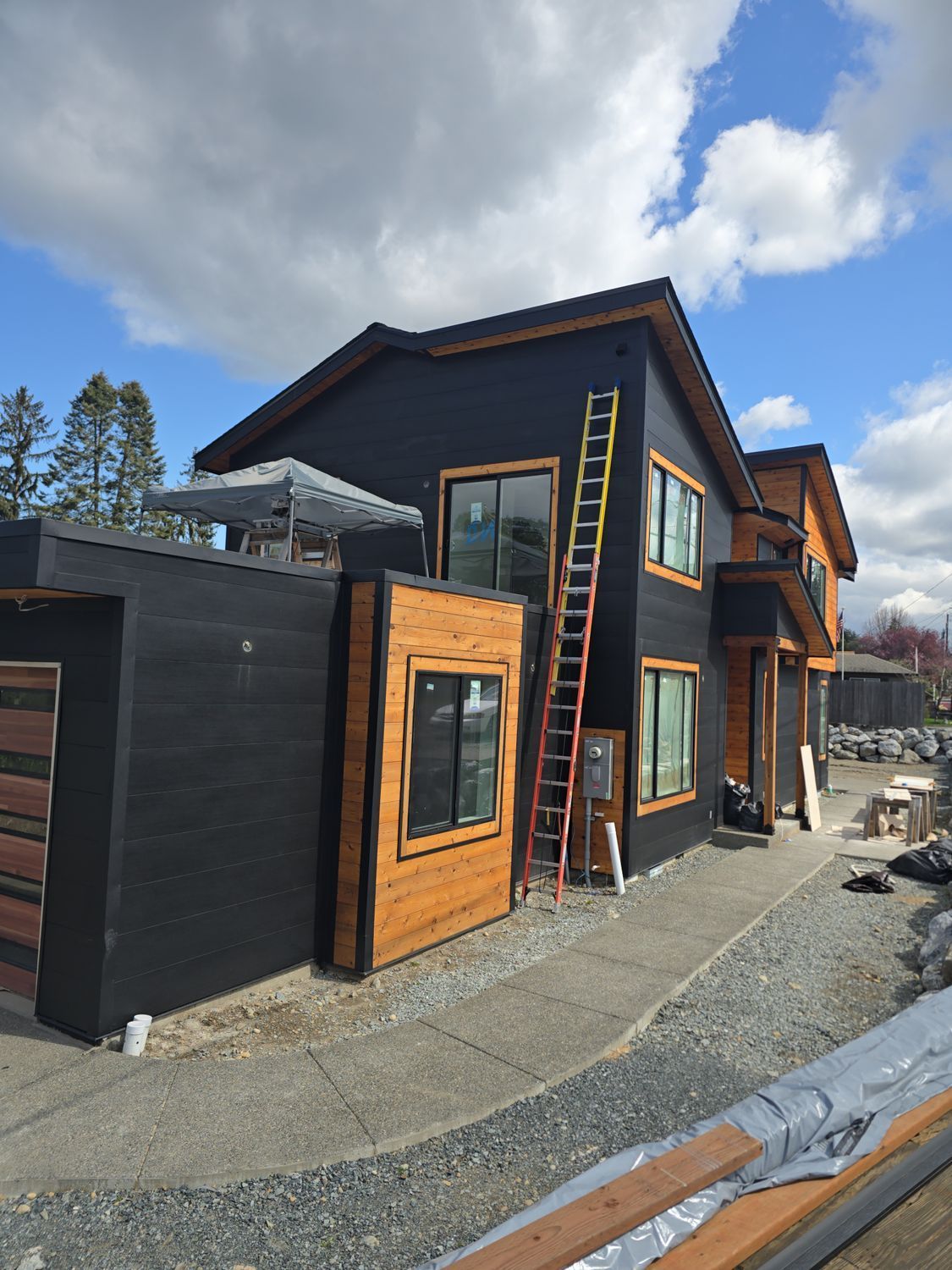 Black and wood-toned two-story house under construction, ladder against the side, cloudy sky above.