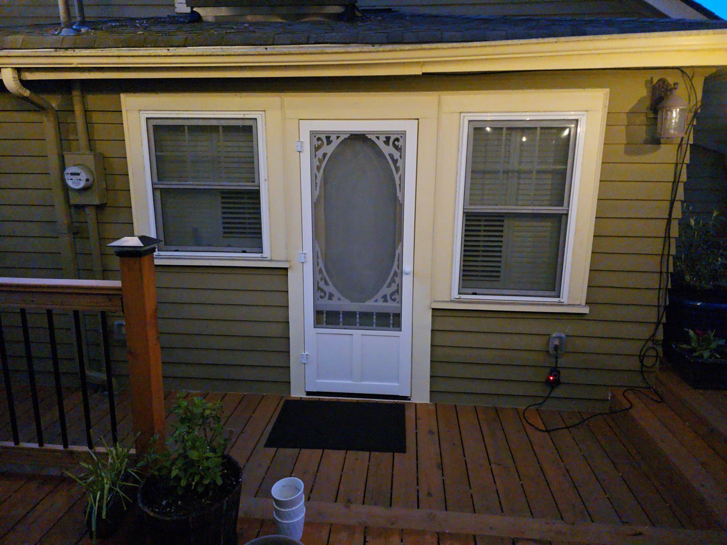 Back door with screen, flanked by windows, on a wooden deck. Olive green siding, white trim, and a black doormat.